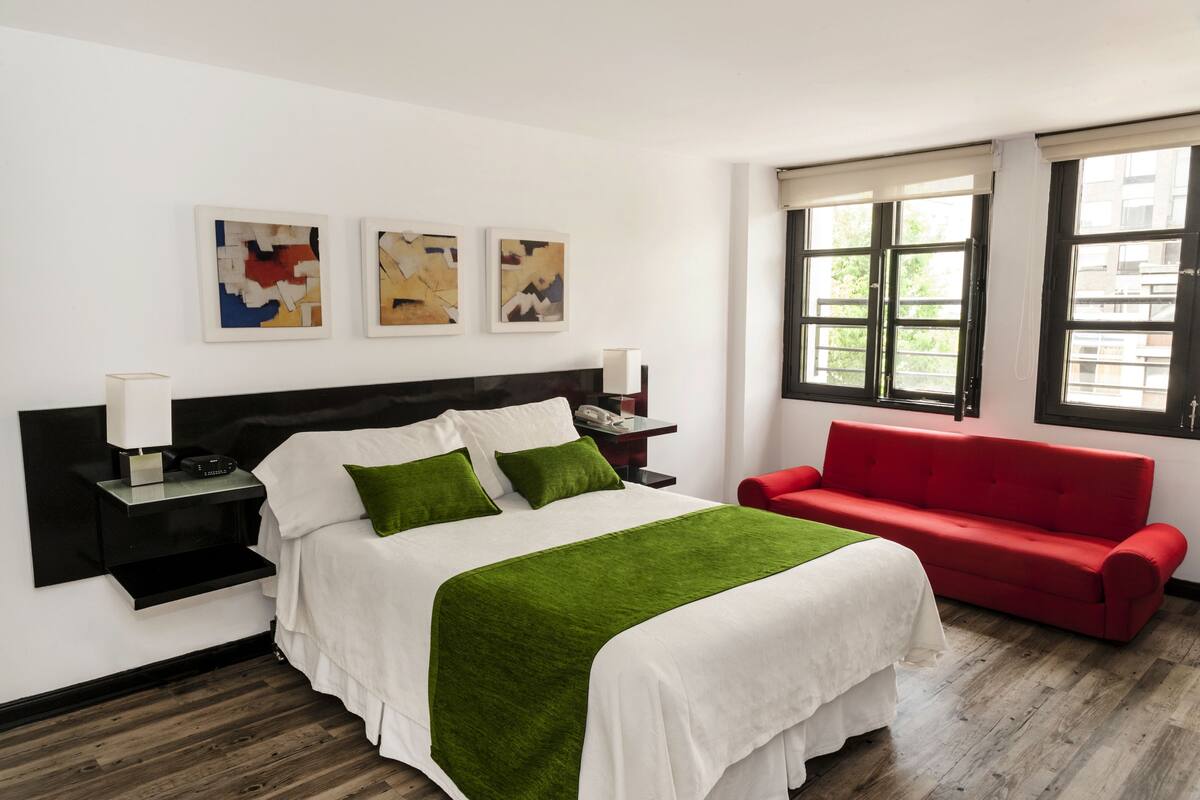 Una camera d'albergo con un letto, un divano rosso, un comodino, una lampada e tre quadri astratti appesi al muro.