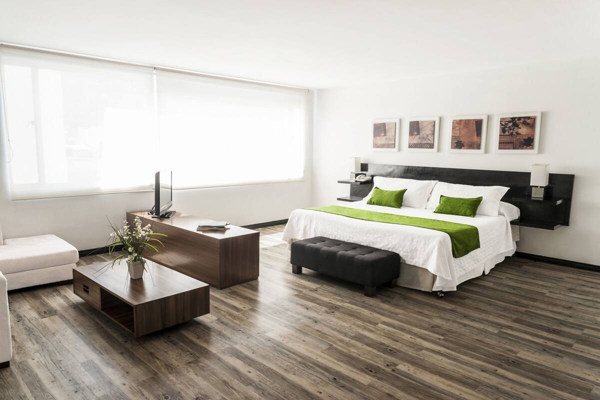 Camera d'albergo moderna con un letto grande, un tavolino in legno, una poltrona bianca e un'ampia finestra con tende.