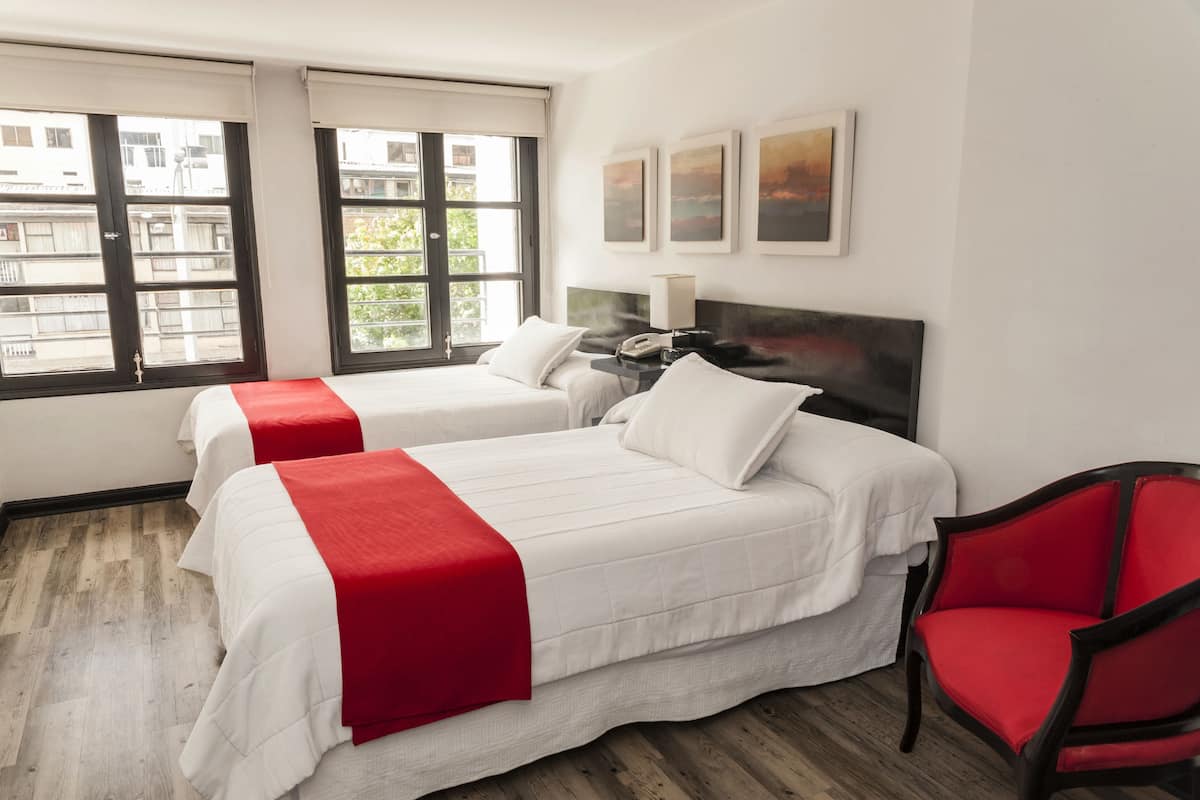 Una camera d'albergo con due letti, una poltrona rossa, un comodino con una lampada e opere d'arte appese al muro.