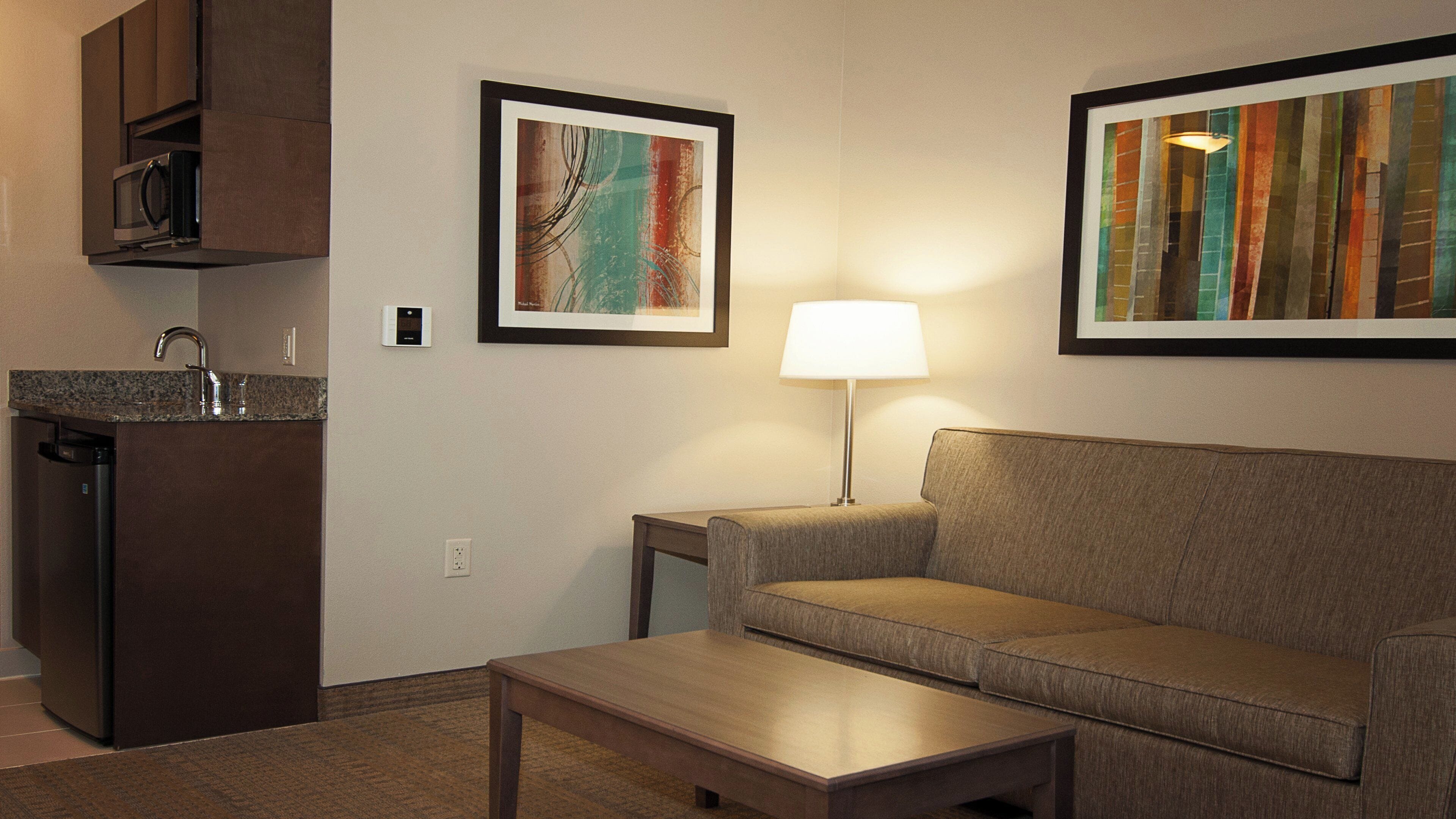 A hotel room with a kitchenette, a sofa, a coffee table, a lamp, and two framed artworks on the wall.
