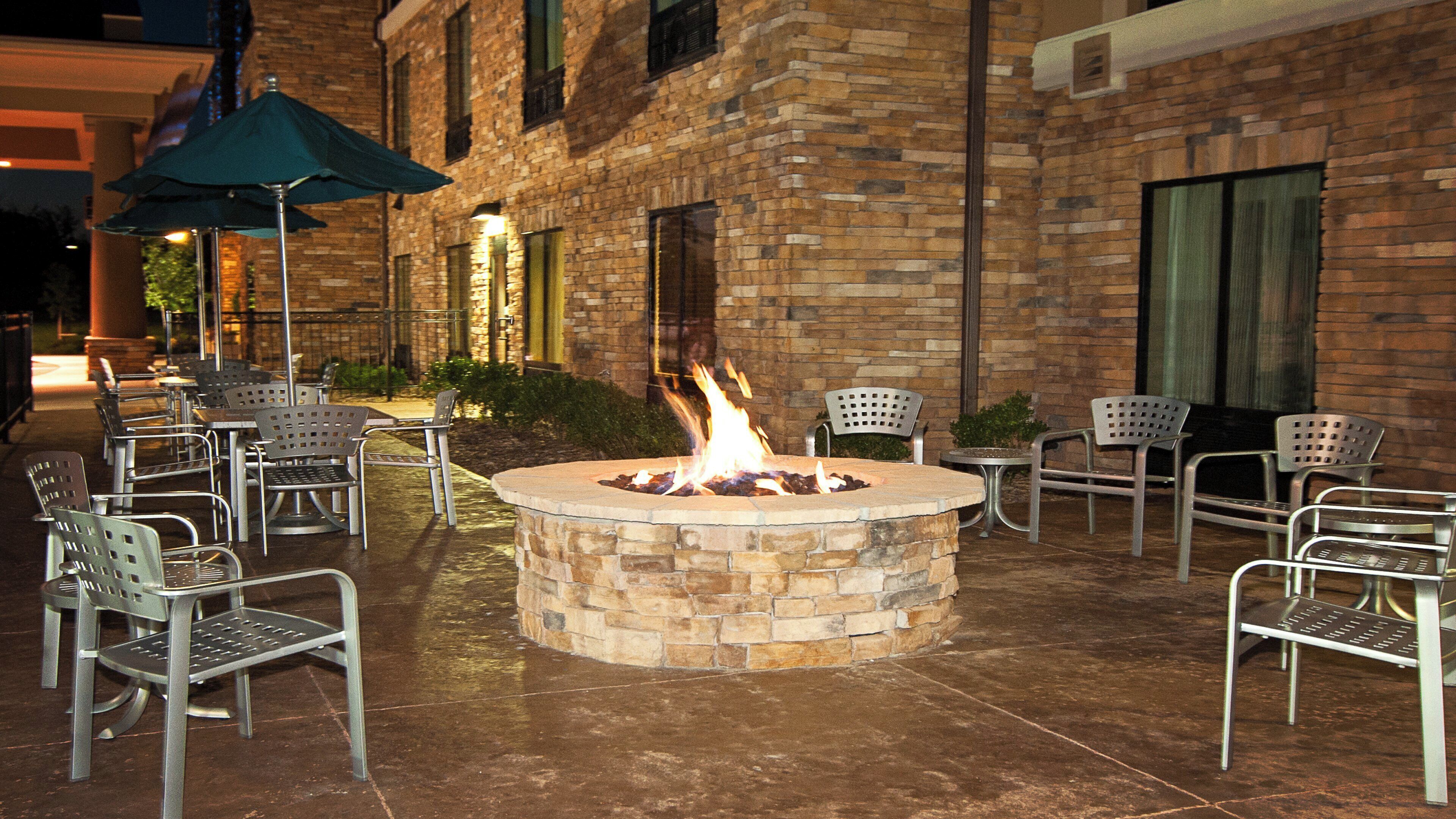 An outdoor seating area with a fire pit surrounded by metal chairs and tables, set against a brick building facade.
