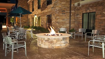 An outdoor seating area with a fire pit surrounded by metal chairs and tables, set against a brick building facade.