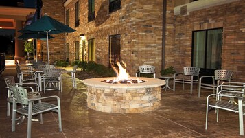 An outdoor seating area with a fire pit surrounded by metal chairs and tables, set against a brick building facade.