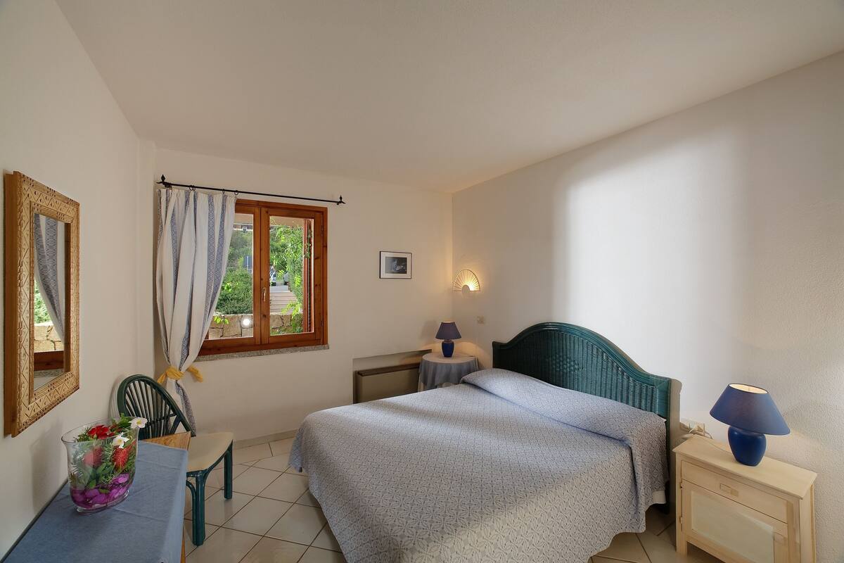 Een slaapkamer met een bed, nachtkastje, lamp, stoel en een raam met gordijnen.