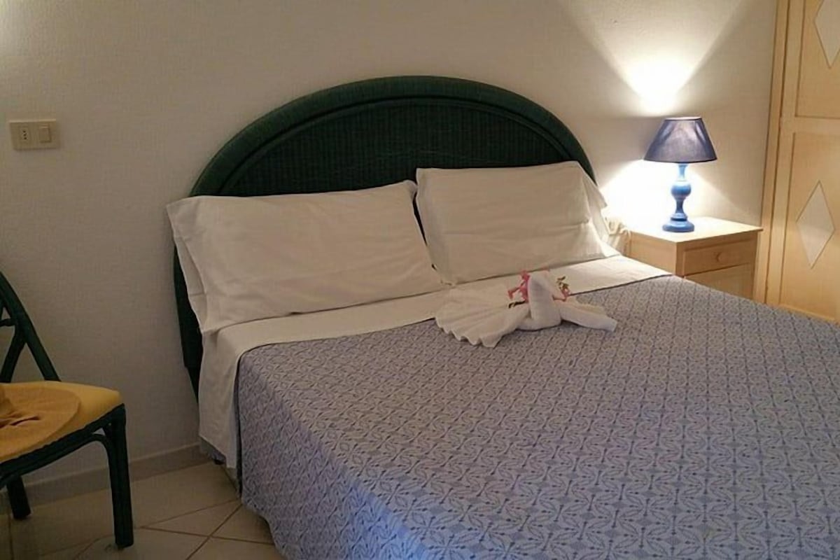 A neatly made bed with a patterned bedspread, a pillow, and a towel. There is a chair and a nightstand with a lamp.