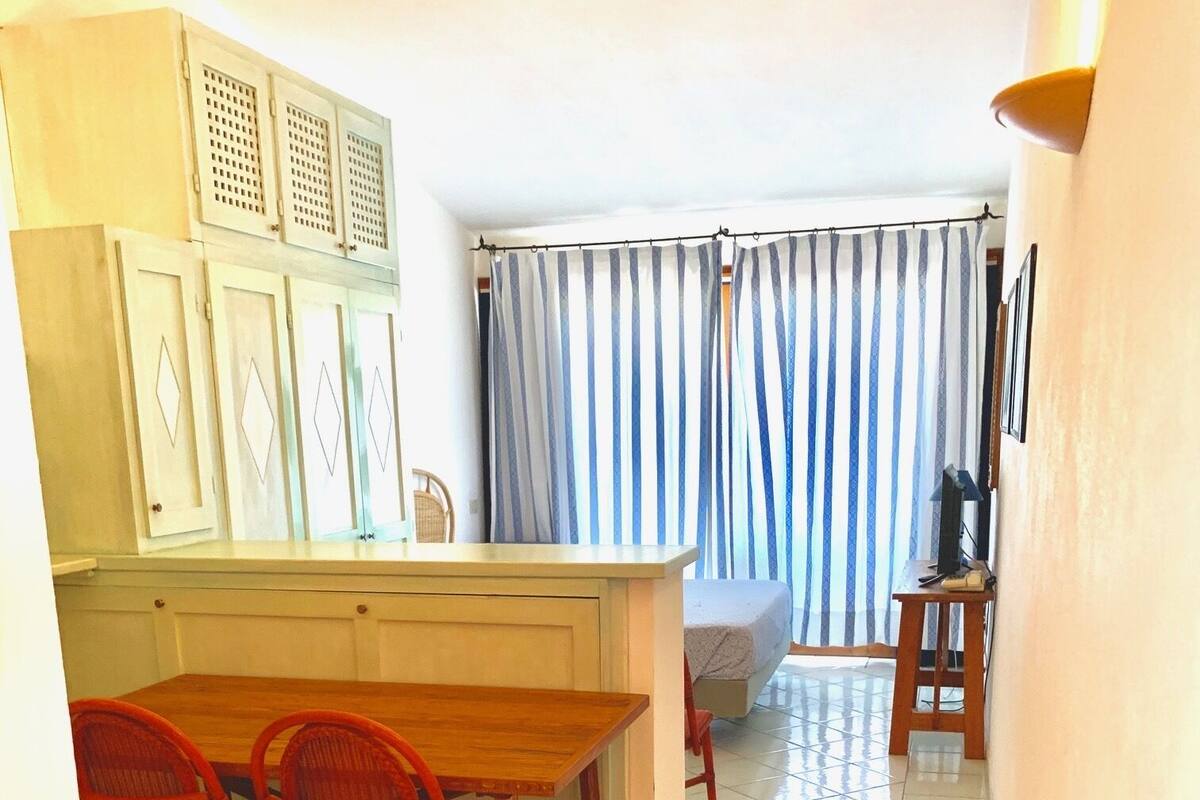 Een kleine keuken met een houten tafel en stoelen, een eethoekje en uitzicht op een slaapkamer door een raam met gordijnen.