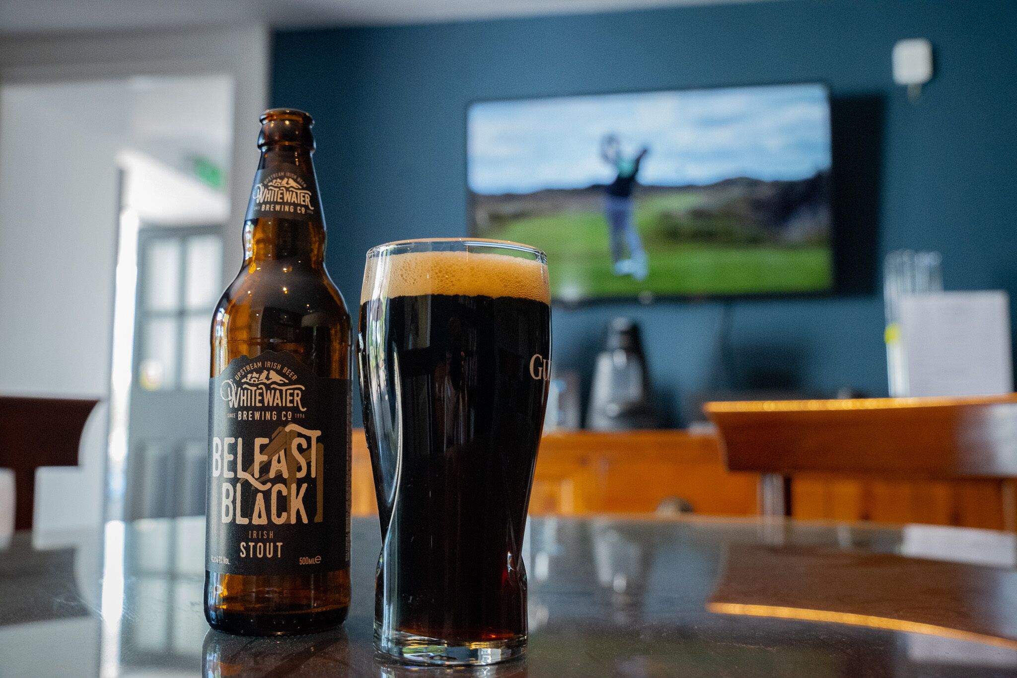 A bottle of Whitewater Brewing Co. Belfast Black Irish Stout and a glass of the same beer on a bar counter with a TV in the background.