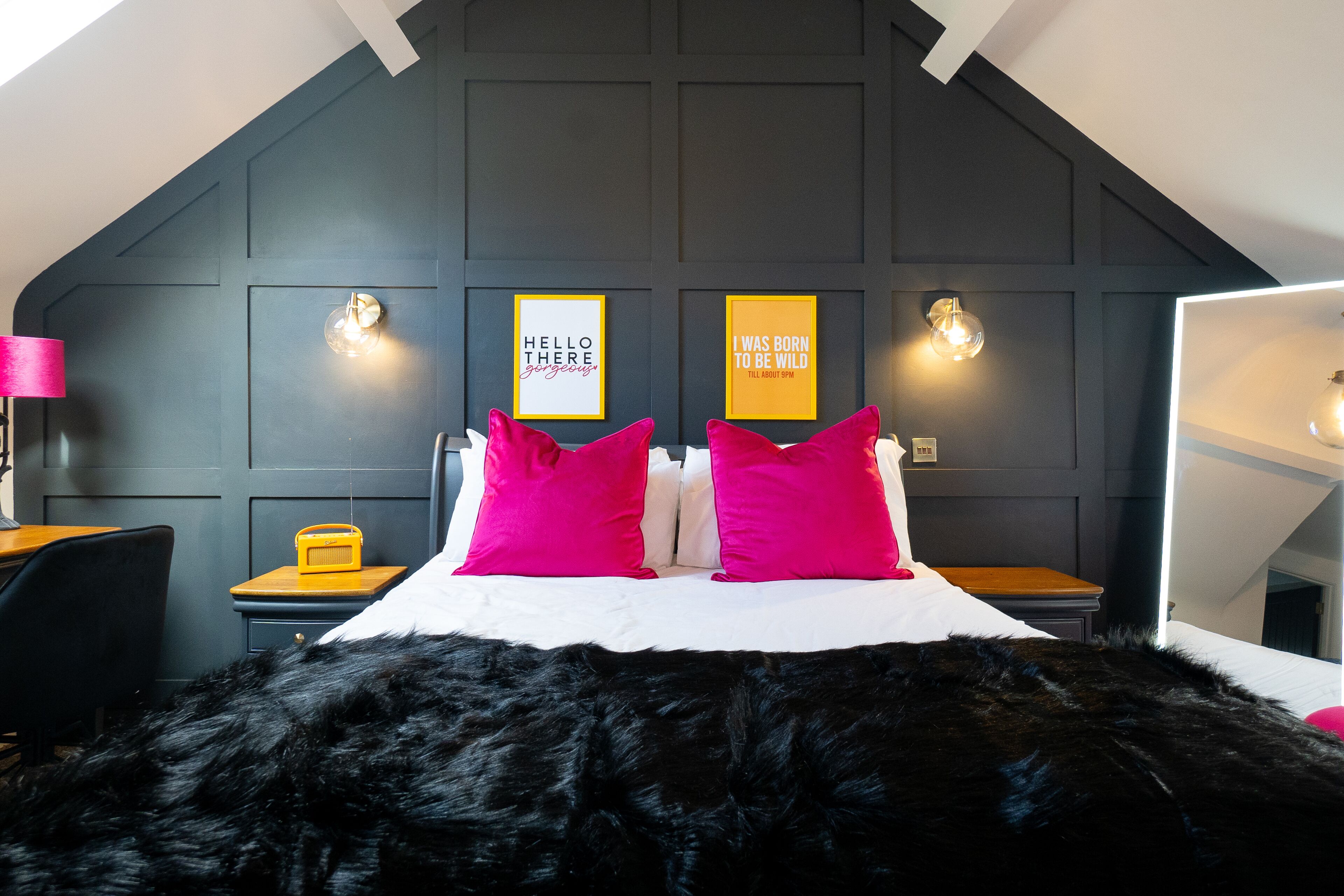 Un dormitorio con un felpudo negro, una cama con dos almohadas rosa brillante, una mesita de noche de madera con una radio amarilla, y dos lámparas de pared con citas motivacionales.