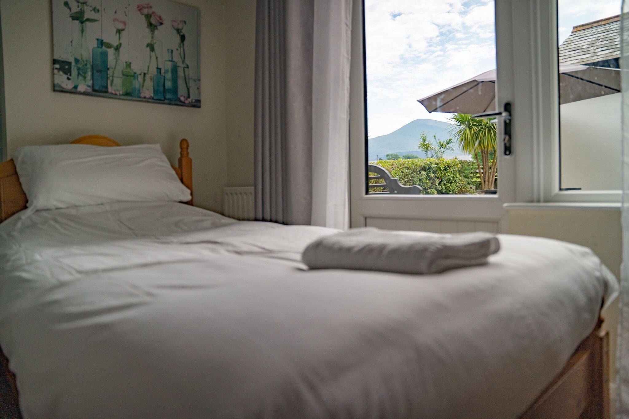 A neatly made bed with white linens, a painting of flowers on the wall, and a view of greenery and mountains through a window.