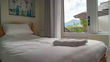 A neatly made bed with white linens, a painting of flowers on the wall, and a view of greenery and mountains through a window.