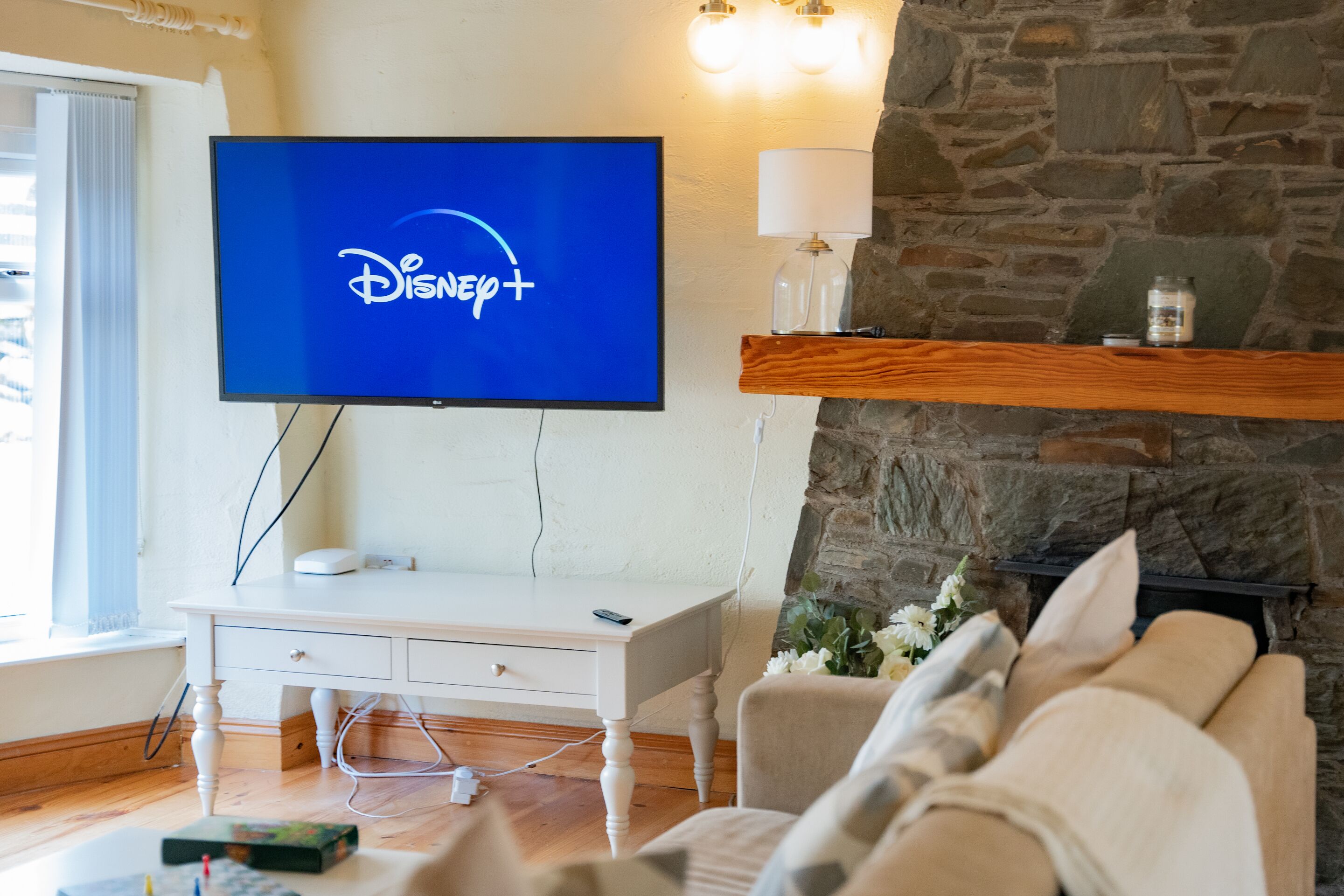 A living room with a flat-screen TV displaying the Disney+ logo, a white desk, a couch, and a stone fireplace.