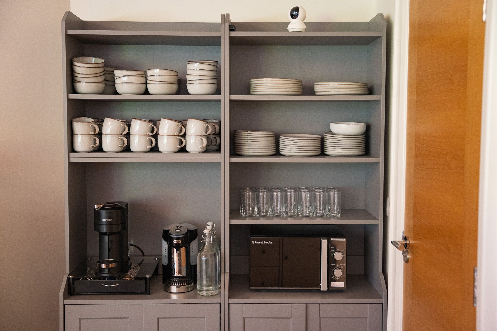 Una cocina con estantes que sostienen platos, tazones y tazas. Hay una cafetera y una botella en la encimera.