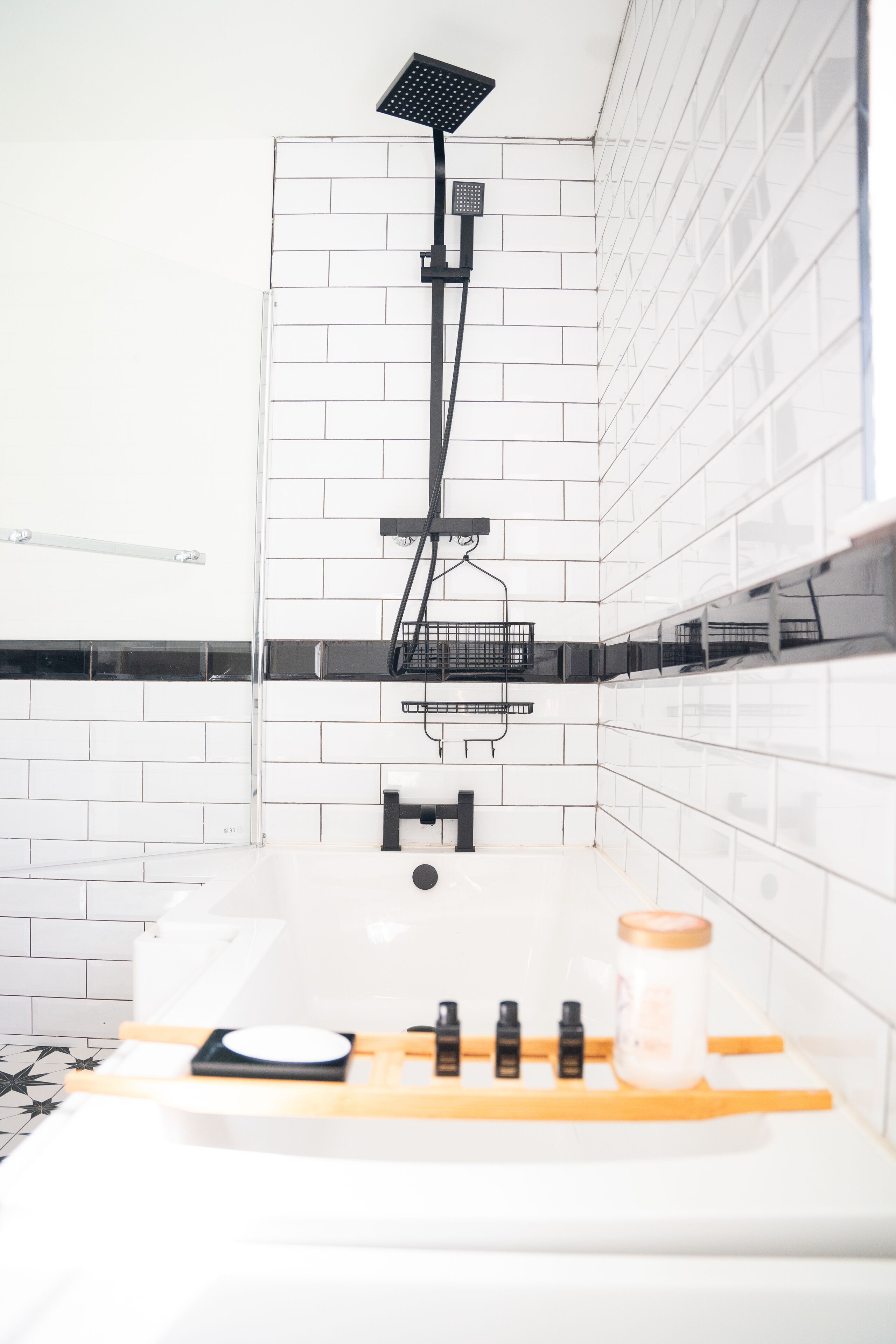 A bathroom with white tiled walls, a black showerhead, a white sink with a wooden tray, and a bottle of shampoo.