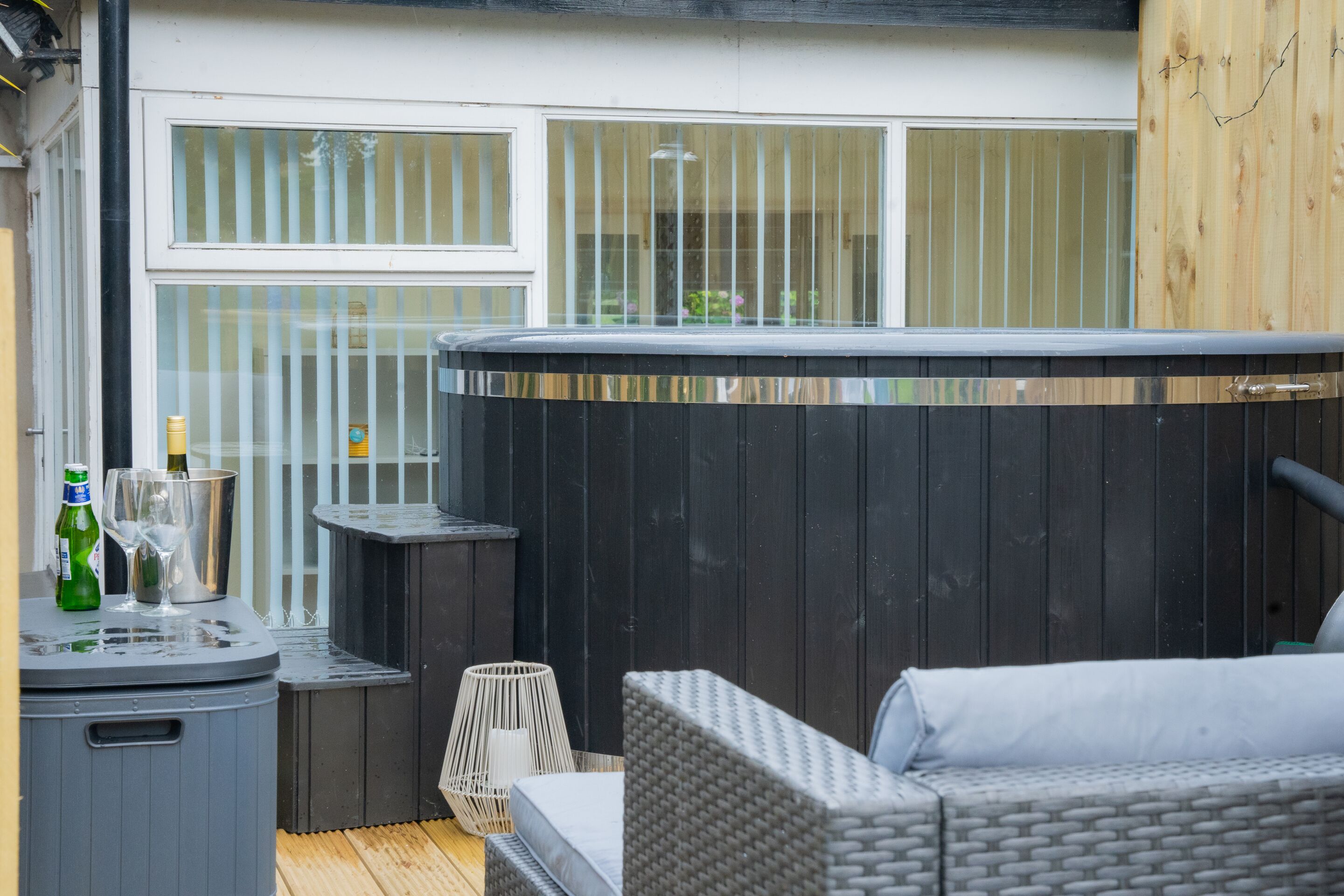 Ein Bereich mit Whirlpool, einer grauen Rattan-Couch, einem kleinen Tisch mit Flaschen und Gläsern sowie einer Holzterrasse.