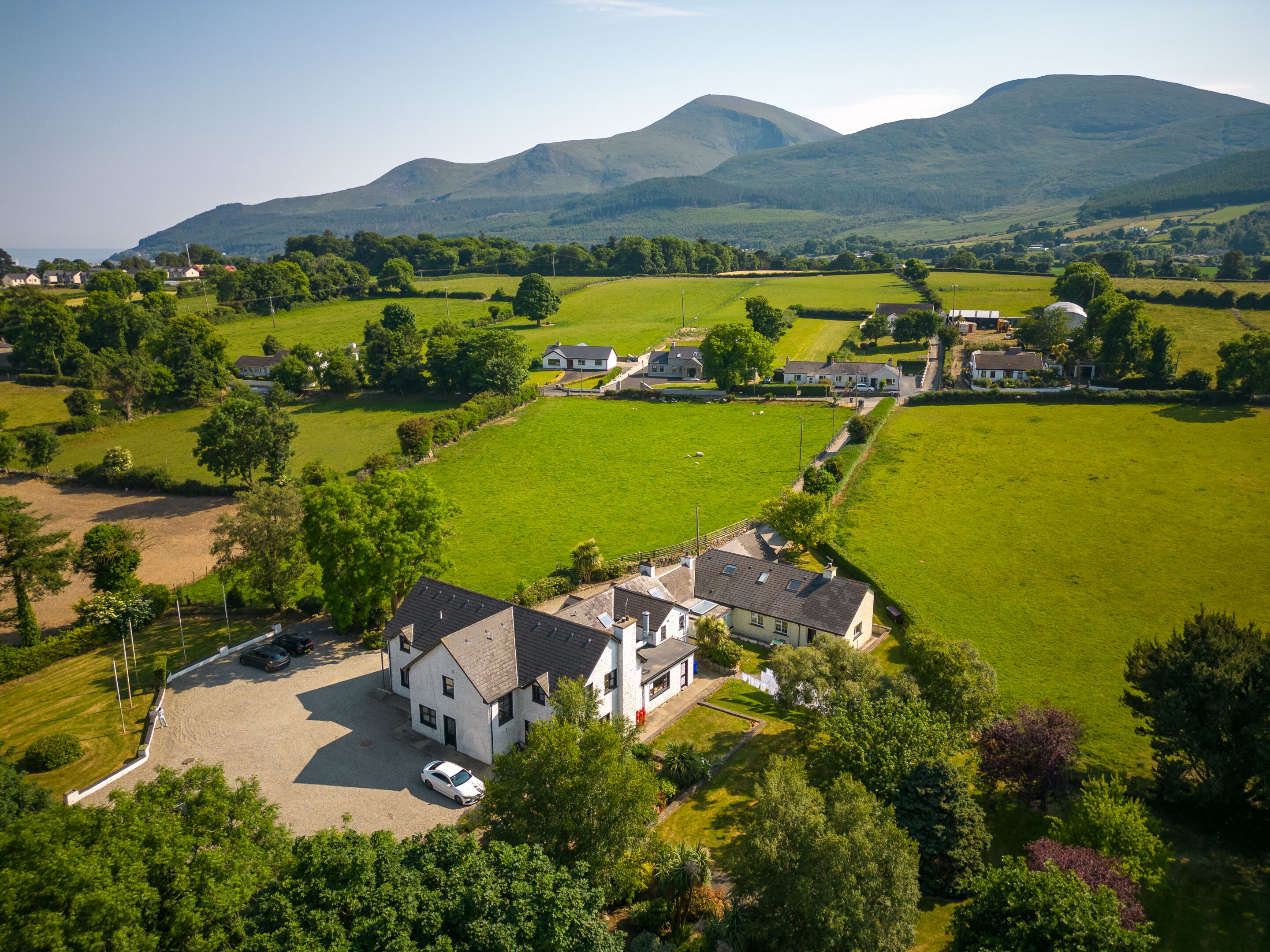 A rural landscape with a large house surrounded by green fields and mountains in the background.