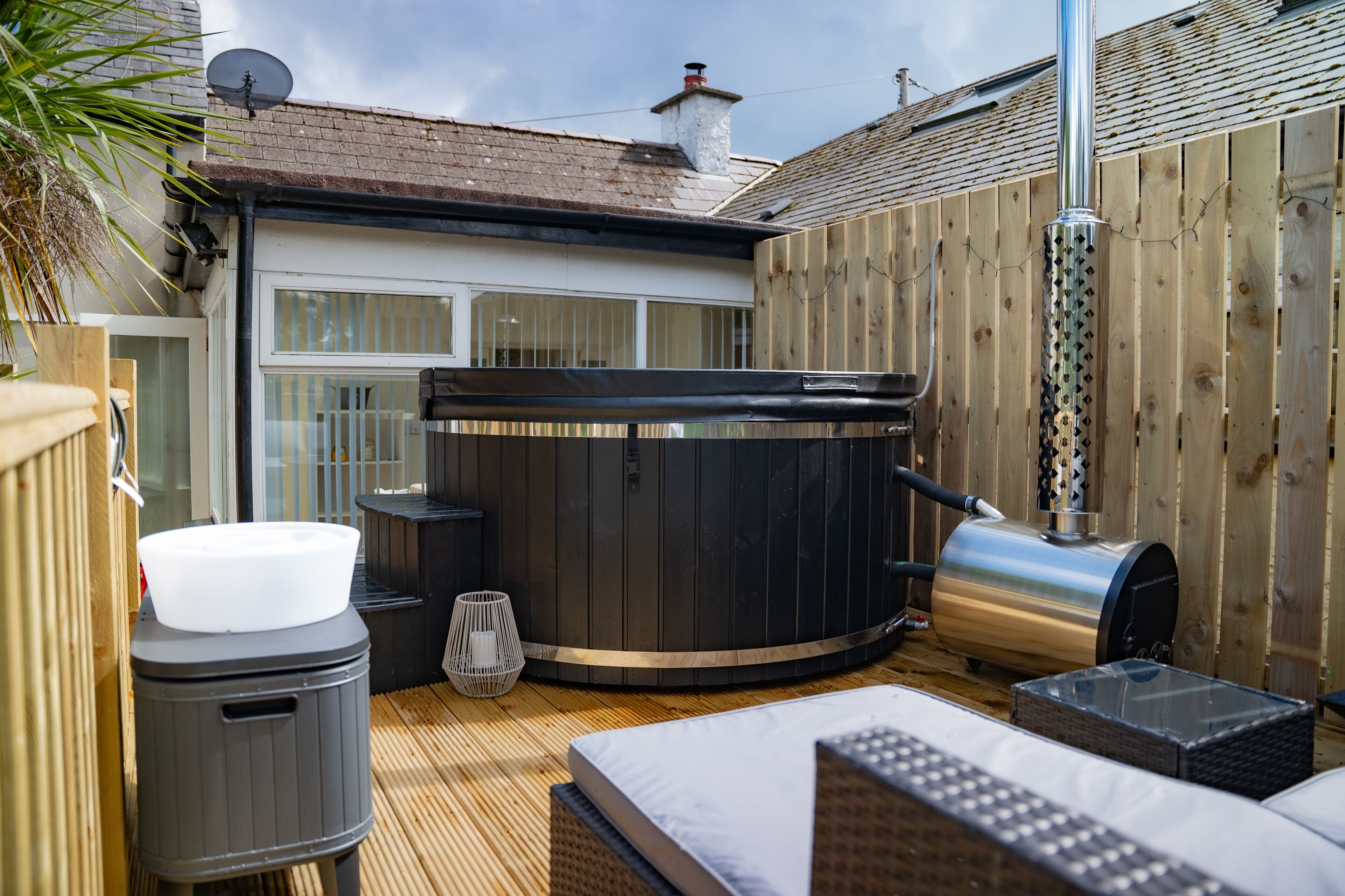 Un bain à remous, un barbecue et une poubelle dans un jardin, avec une clôture en bois et une maison en arrière-plan.