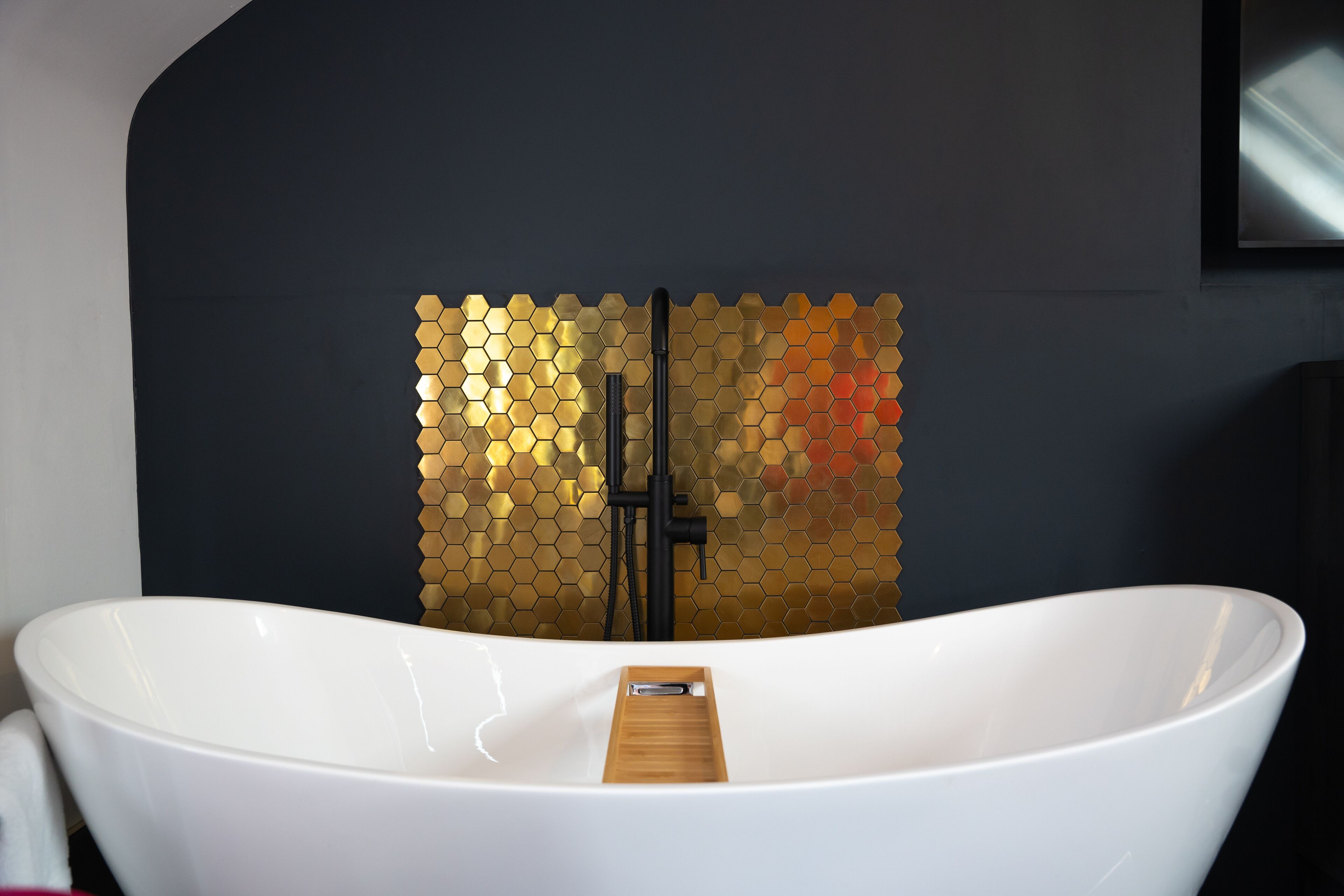 Ein modernes Badezimmer mit einer weißen, freistehenden Badewanne, einer Wand mit goldfarbenen, sechseckigen Mosaiken und einer schwarzen Armatur.