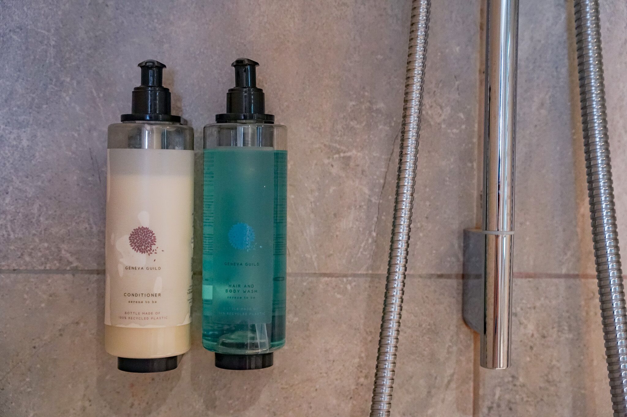 Two bottles of Geneva Guild hair and body wash next to a shower hose.