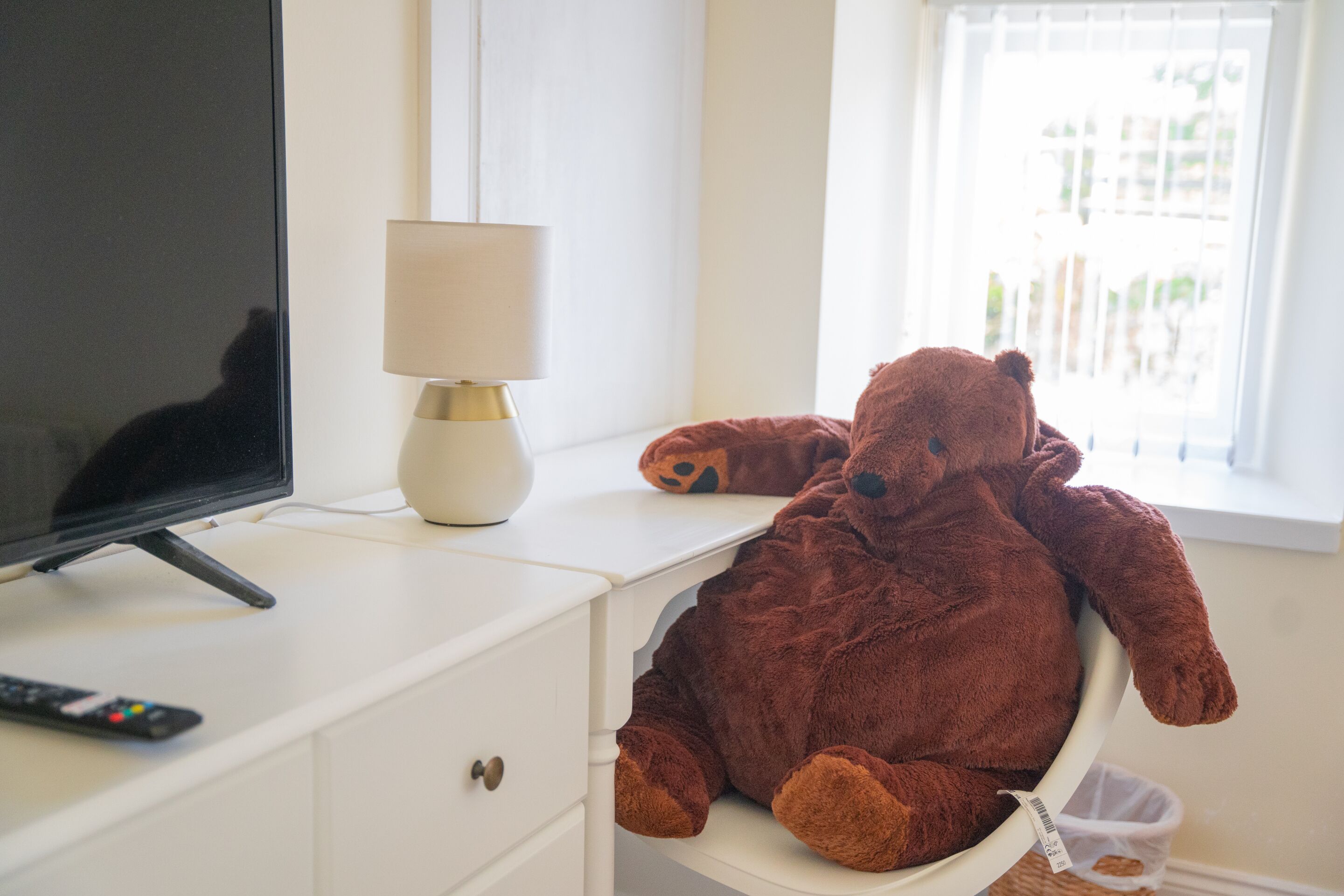 A room with a television on a white cabinet, a lamp, and a large teddy bear on a chair.