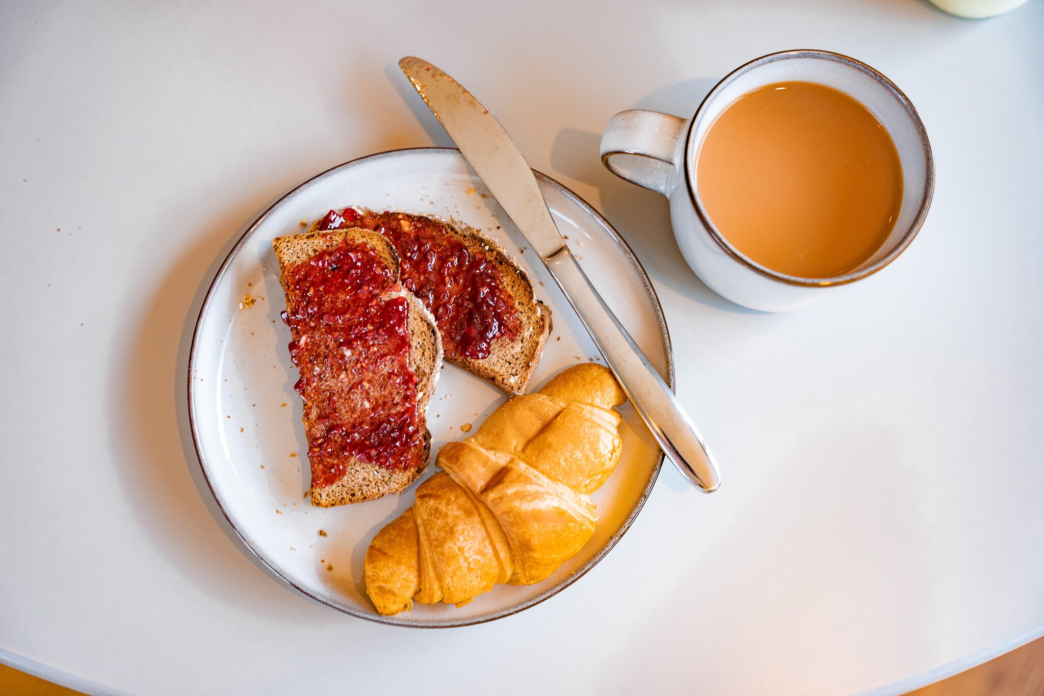 En tallrik med två skivor rostat bröd och en croissant, en kniv och en kopp kaffe på ett vitt bord.