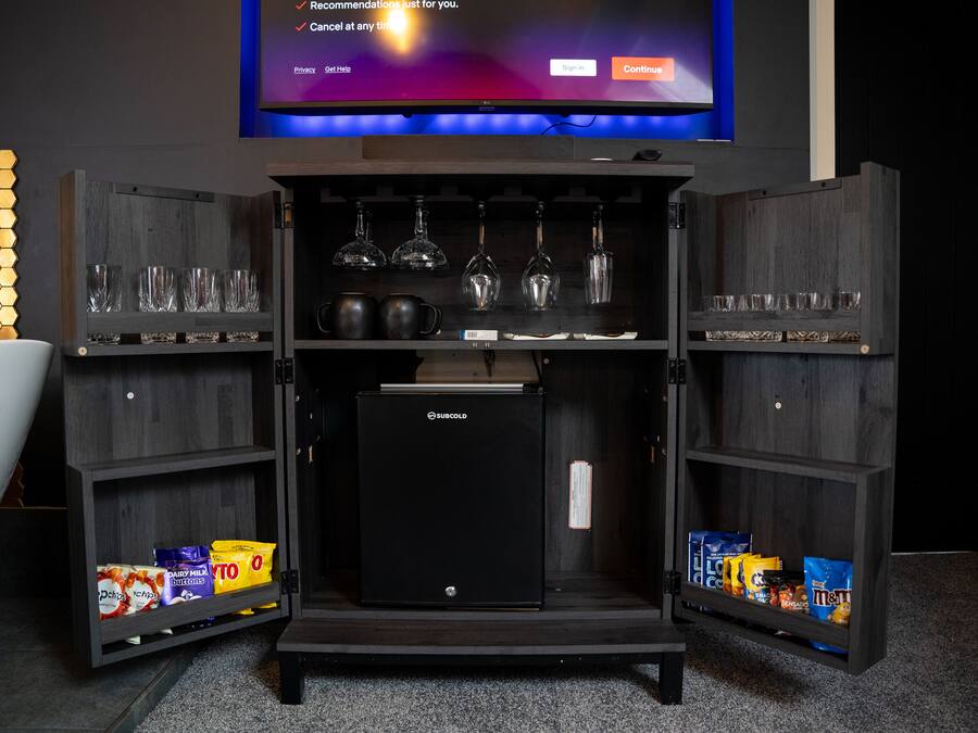 A compact minibar with a refrigerator, shelves with glasses and bottles, and snack packages.