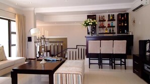 A modern living room with a sofa, a dining table, a bar area with shelves of bottles, and a television. - Sanchia Luxury Guesthouse (Durban North)