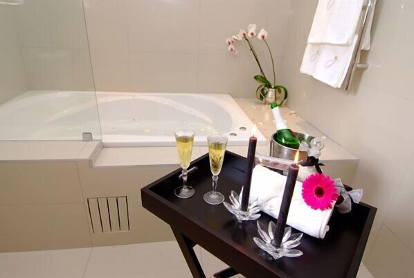 A bathroom with a bathtub, a small table with two glasses of champagne, a vase with flowers, and a towel rack.