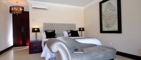 A modern bedroom with a large bed, two bedside tables, a chandelier, and a framed leaf artwork on the wall.
