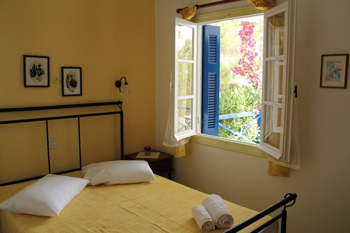 A bed with white pillows, a small bedside table with a lamp, and a window with blue shutters.