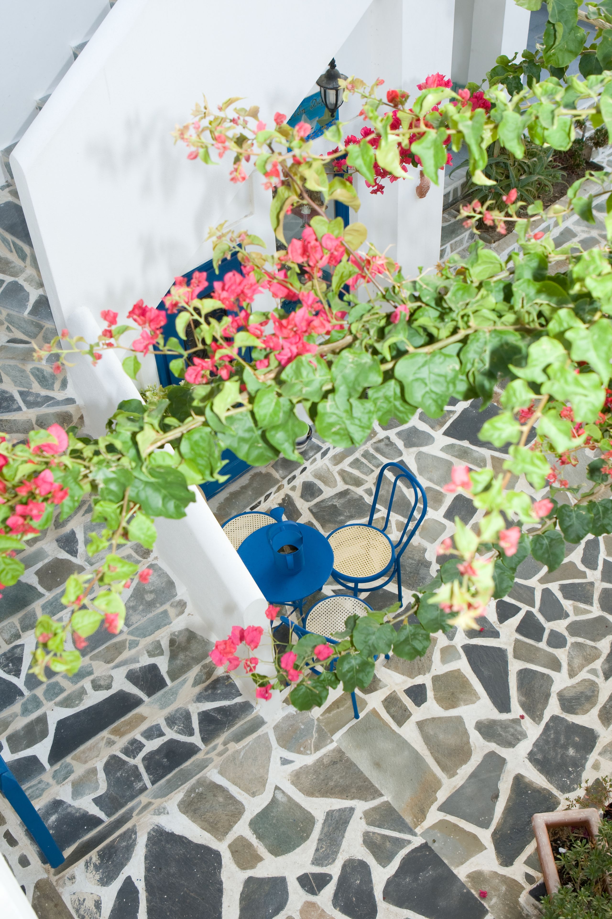 Un patio con suelo de piedra, una silla azul y una mesa blanca, rodeado de follaje verde y flores rosas.