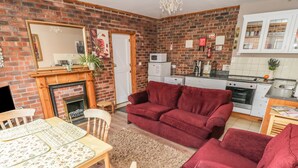 A cozy living room with a brick fireplace, a red sofa, a dining table with chairs, and a kitchen area with cabinets and appliances. - Church View B&B and Holiday Cottages (York)