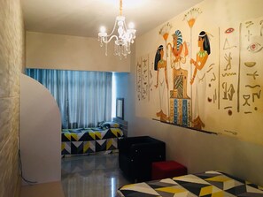 A hotel room with a bed, a red ottoman, a black chair, a chandelier, and a wall with Egyptian hieroglyphs. - Hotel das Palmas (Florianópolis)