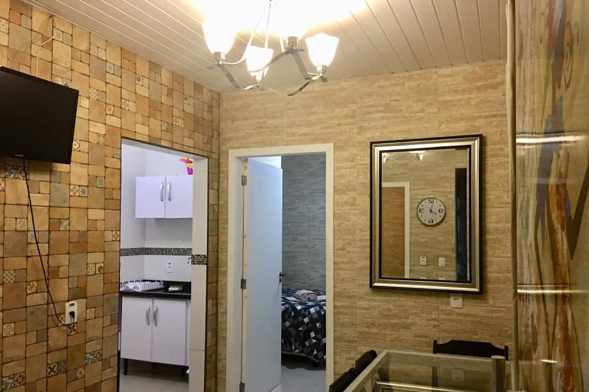 Un ambiente compatto con angolo cottura, zona pranzo con tavolo in vetro e camera da letto con letto e orologio a parete.