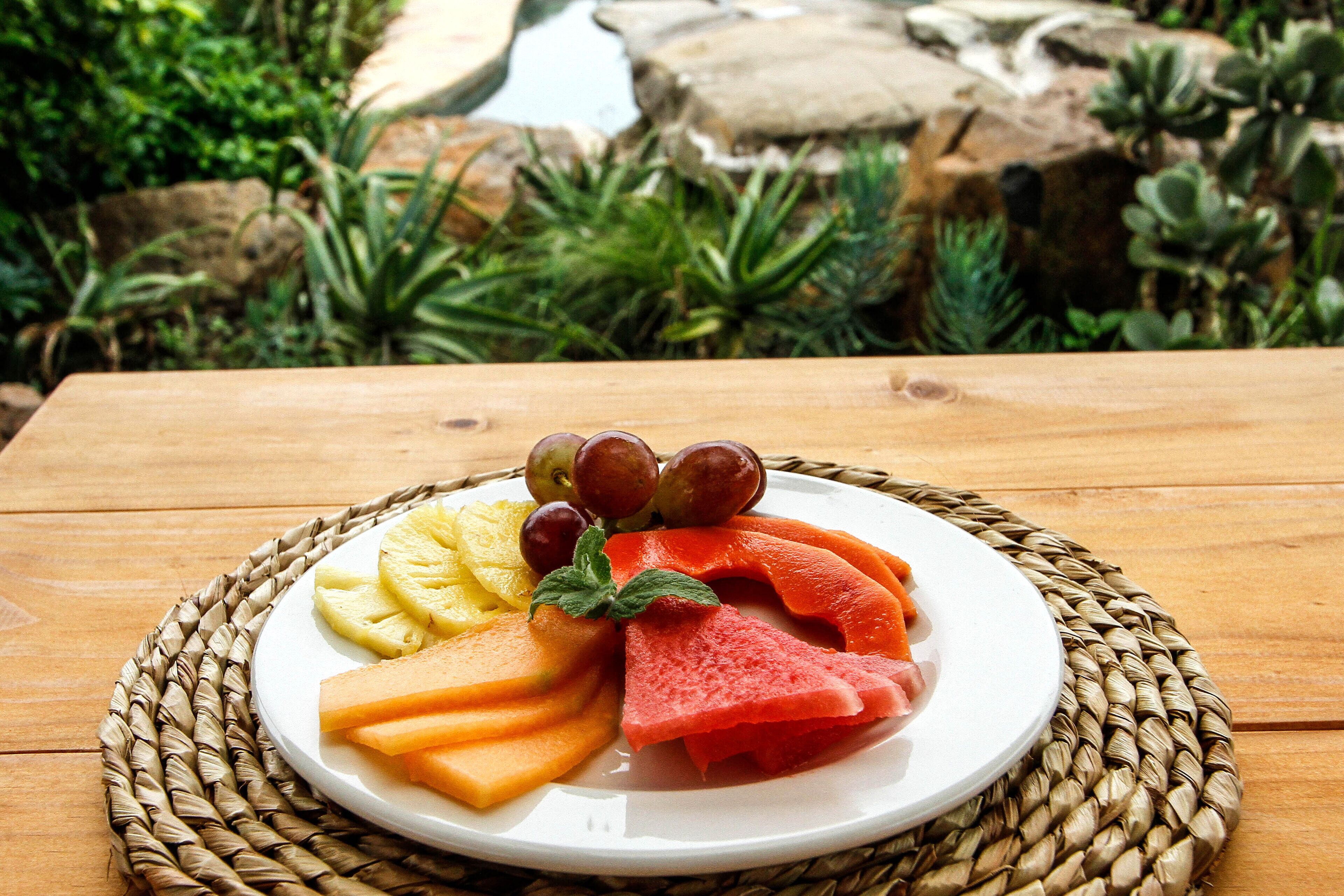 A plate of fruit on a wooden table with a woven placemat, set against a backdrop of rocks and greenery.