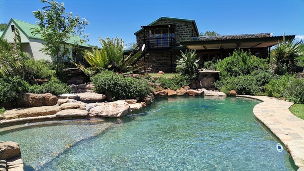 A clear swimming pool with a stone border, surrounded by rocks and greenery, and a stone building in the background. - SpionKop Lodge (Ladysmith)