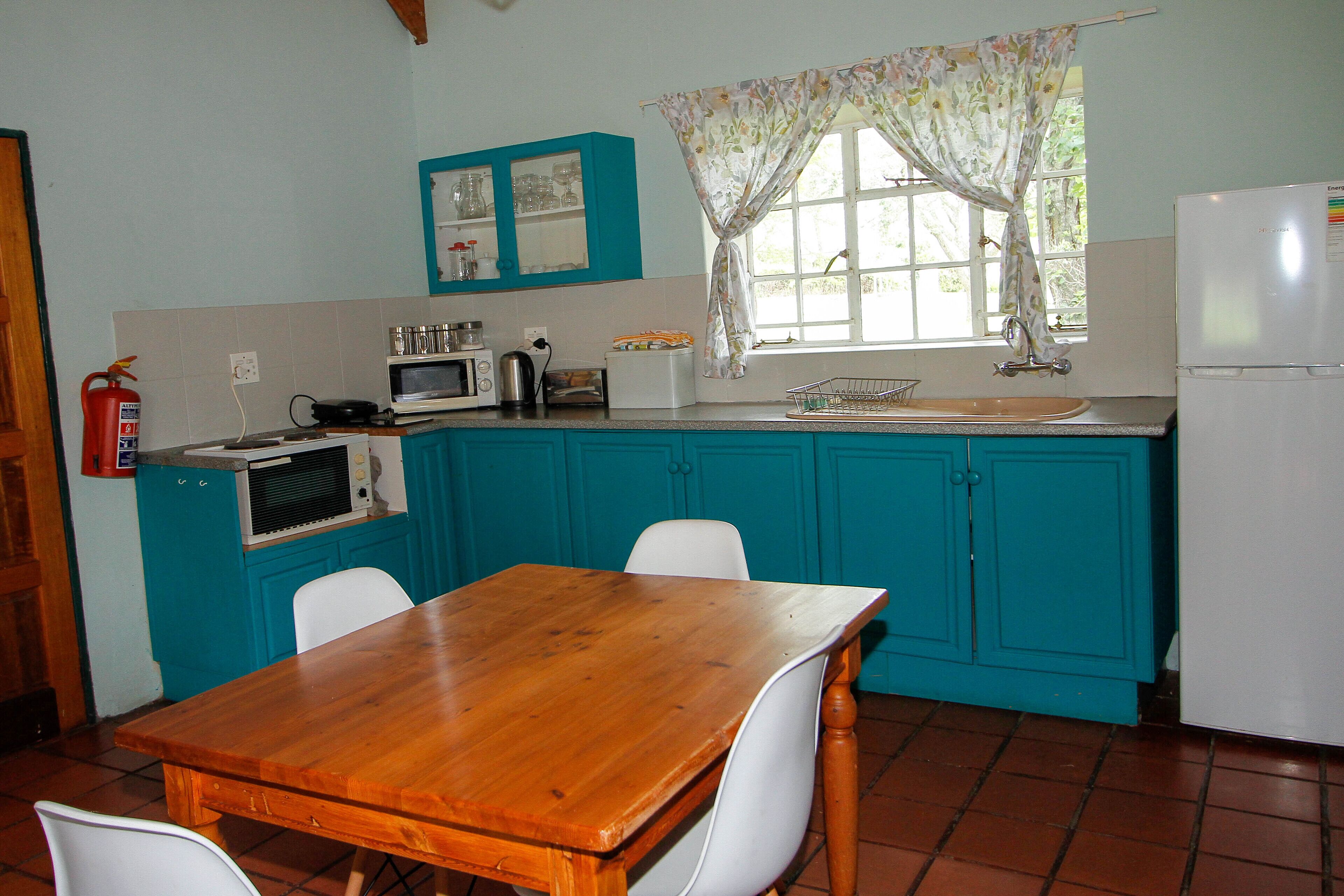 Una cocina con armarios azul turquesa, una mesa de madera, sillas blancas, un microondas, un hervidor y un refrigerador.