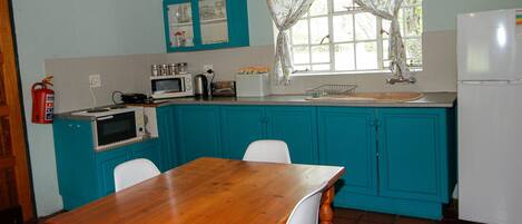 Una cocina con armarios azul turquesa, una mesa de madera, sillas blancas, un microondas, un hervidor y un refrigerador.