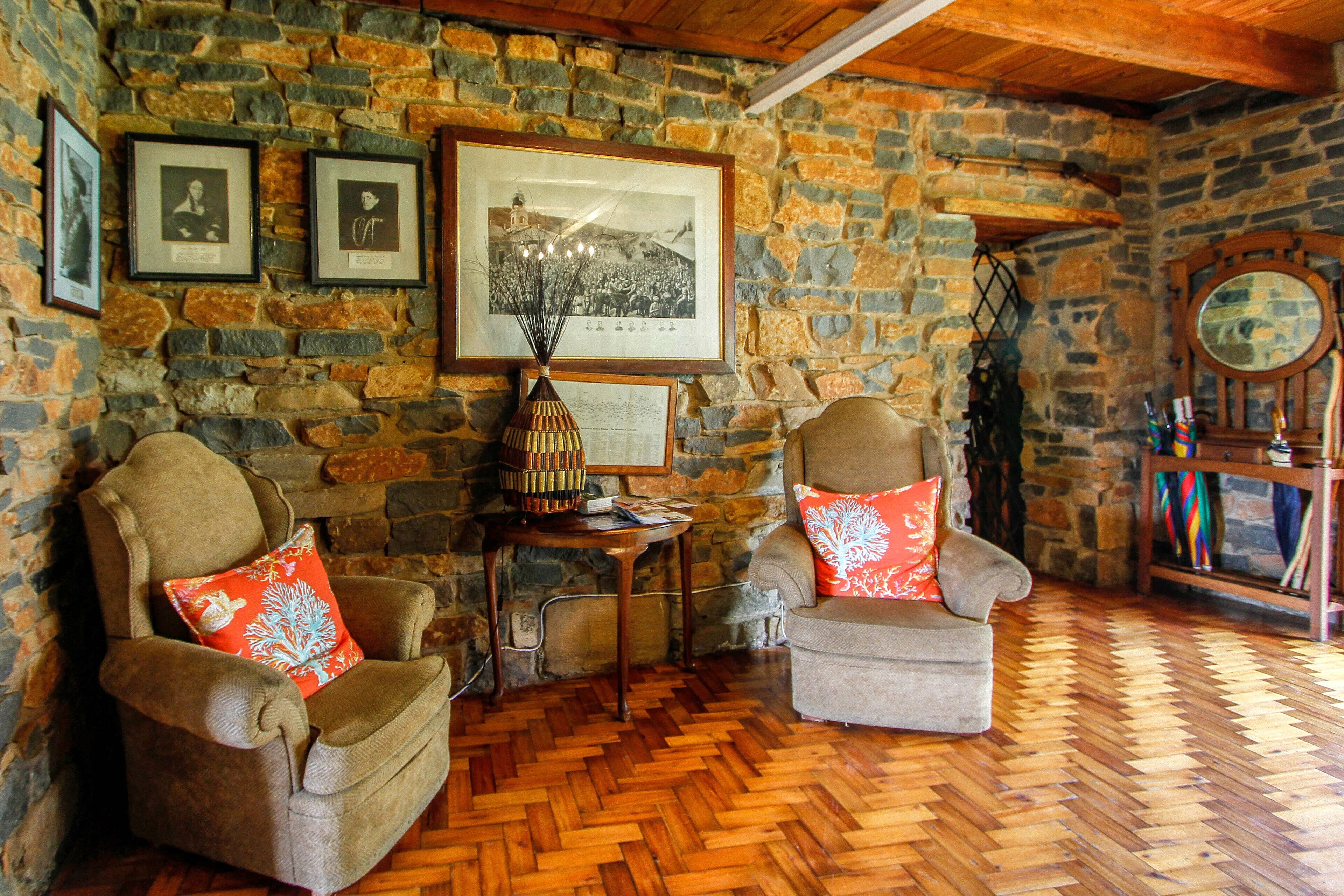 A cozy room with stone walls, wooden flooring, two armchairs with decorative pillows, a small table with a vase, and framed pictures on the wall.