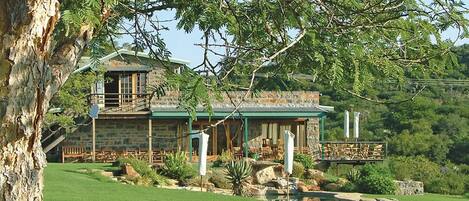 Una casa de piedra con techo verde, rodeada de un césped bien cuidado y un árbol grande.