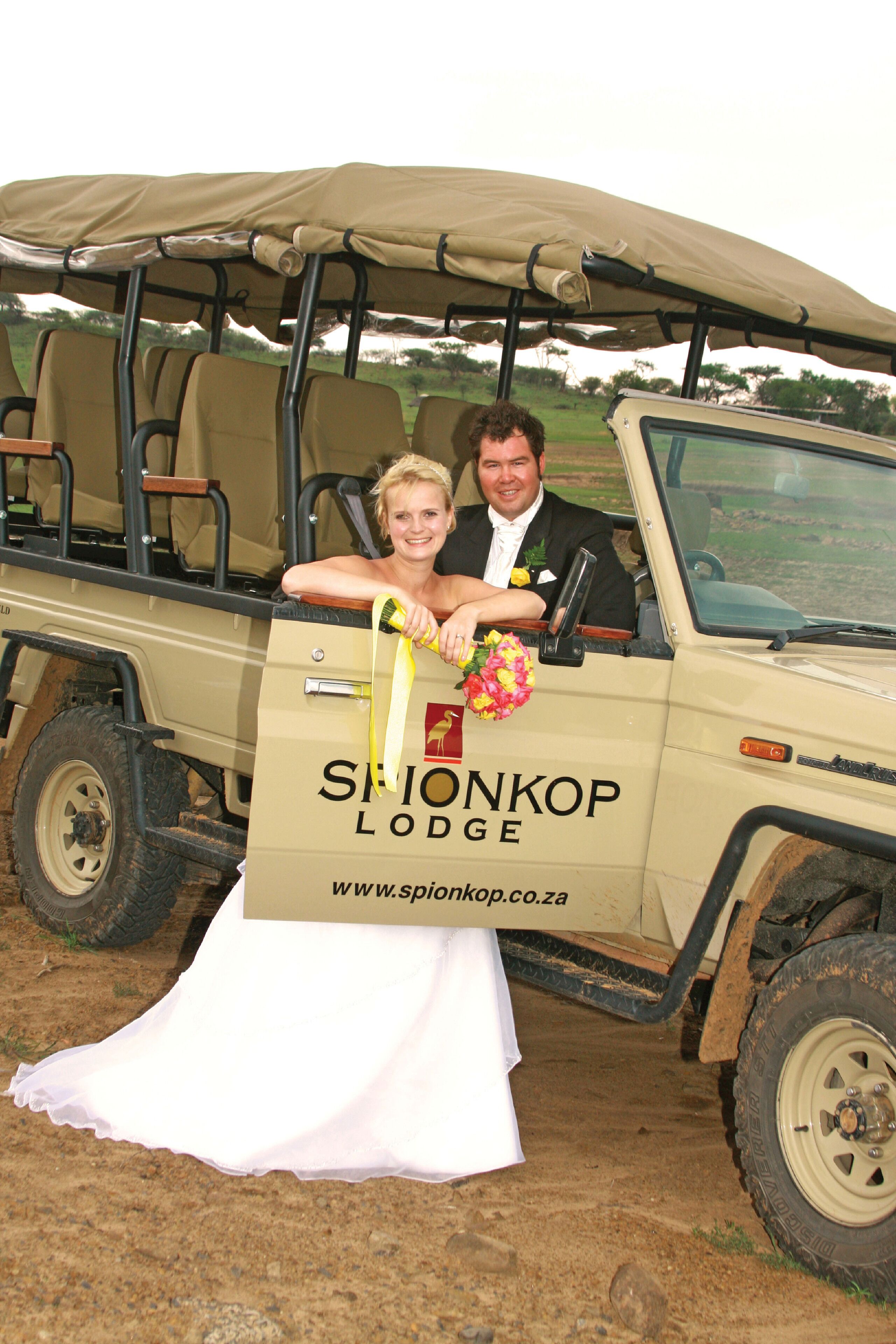 Ein Brautpaar in einem Safarifahrzeug, die Braut trägt ein Hochzeitskleid und einen Blumenstrauß, und ein Schild für Spionkop Lodge.