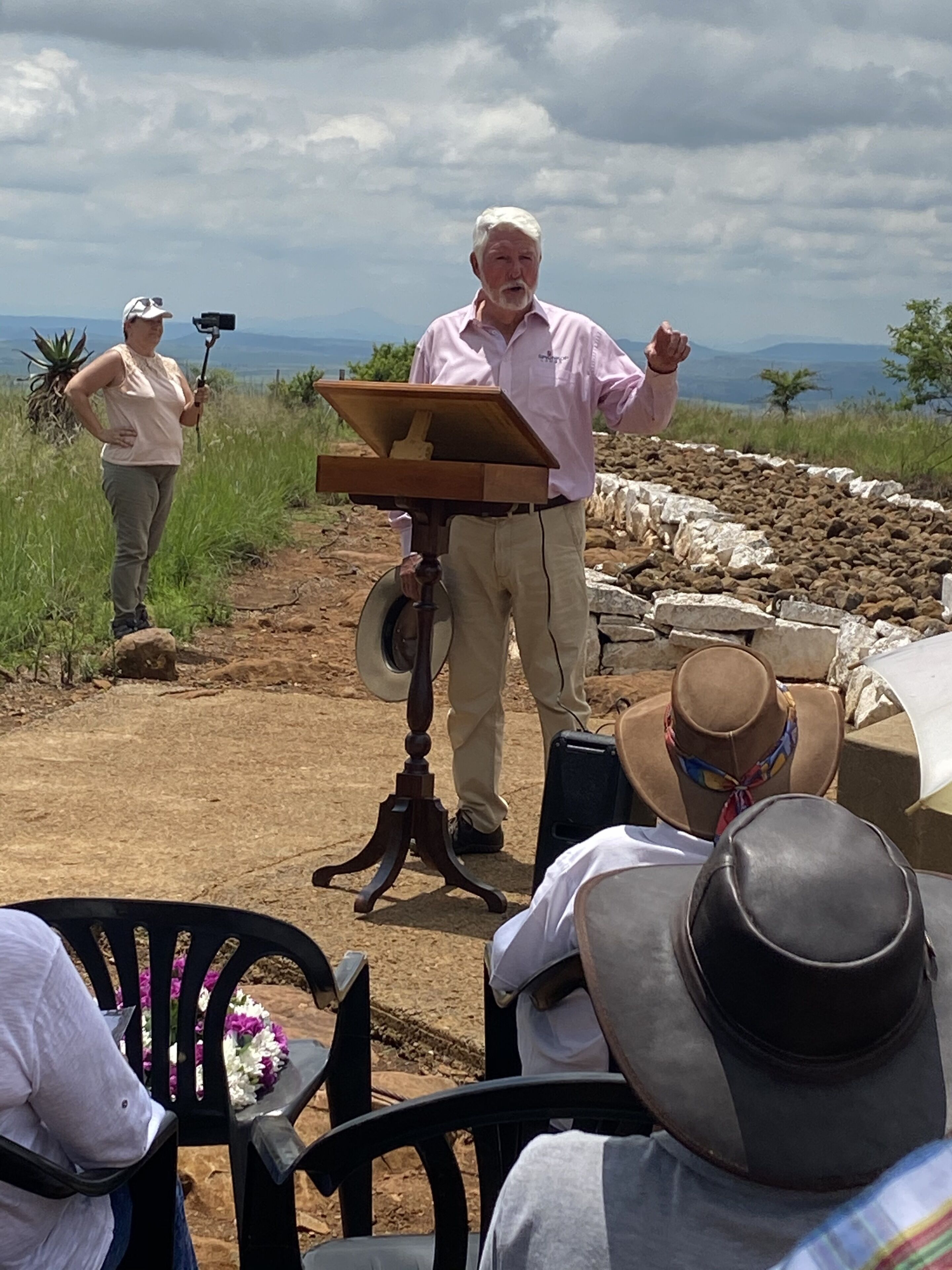 A man speaking at a podium outdoors with a scenic view in the background.