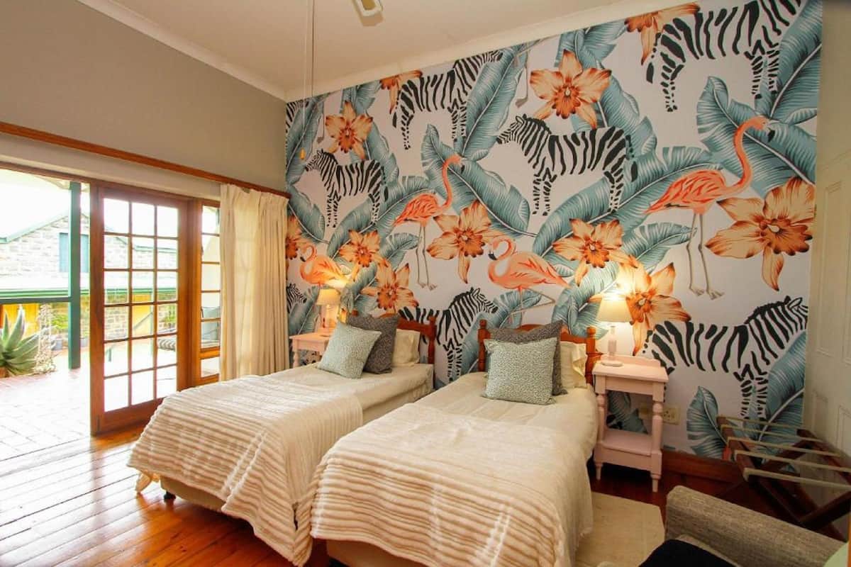 Quarto com duas camas, uma janela grande com cortinas e um mural temático tropical.