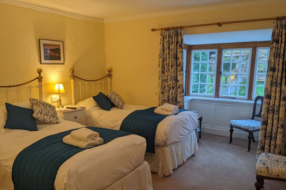 Habitación de hotel con dos camas, una silla, una ventana con cortinas, una mesita de noche con lámpara y un cuadro en la pared.