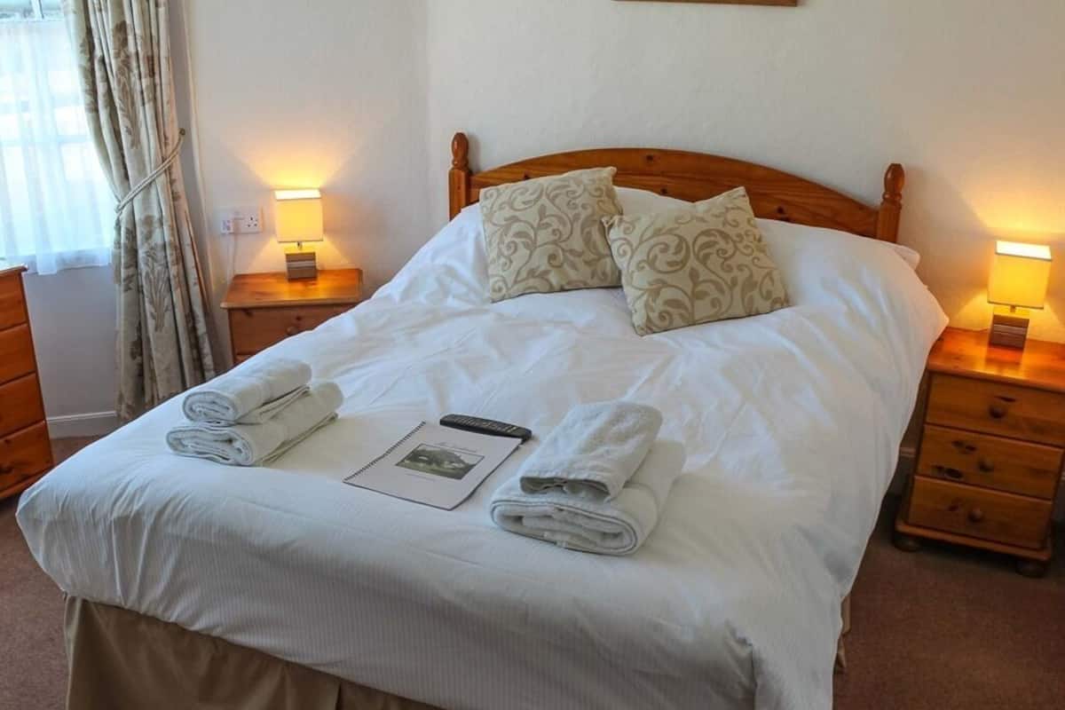 Una cama bien hecha con dos almohadas, dos lámparas de noche, una mesita de noche con lámpara y una toalla enrollada sobre la cama.