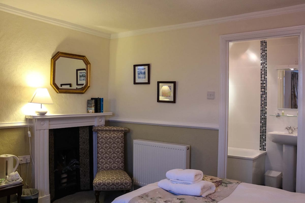 Habitación de hotel con cama, una silla, chimenea, radiador, baño con lavamanos y espejo, y cuadros en la pared.