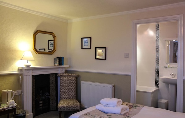 A hotel room with a bed, a chair, a fireplace, a radiator, a bathroom with a sink and mirror, and framed pictures on the wall.