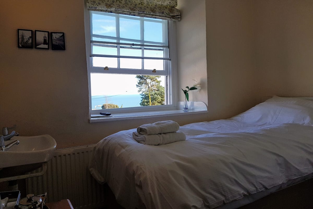 Un dormitorio con un ventanal grande, una mesita de noche con un florero y una cama bien tendida.