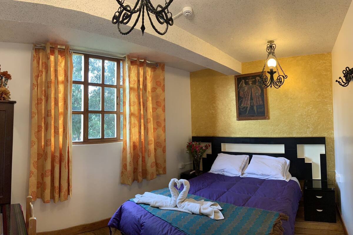 A bedroom with a bed, a chandelier, a window with curtains, a wall-mounted picture, and a nightstand.
