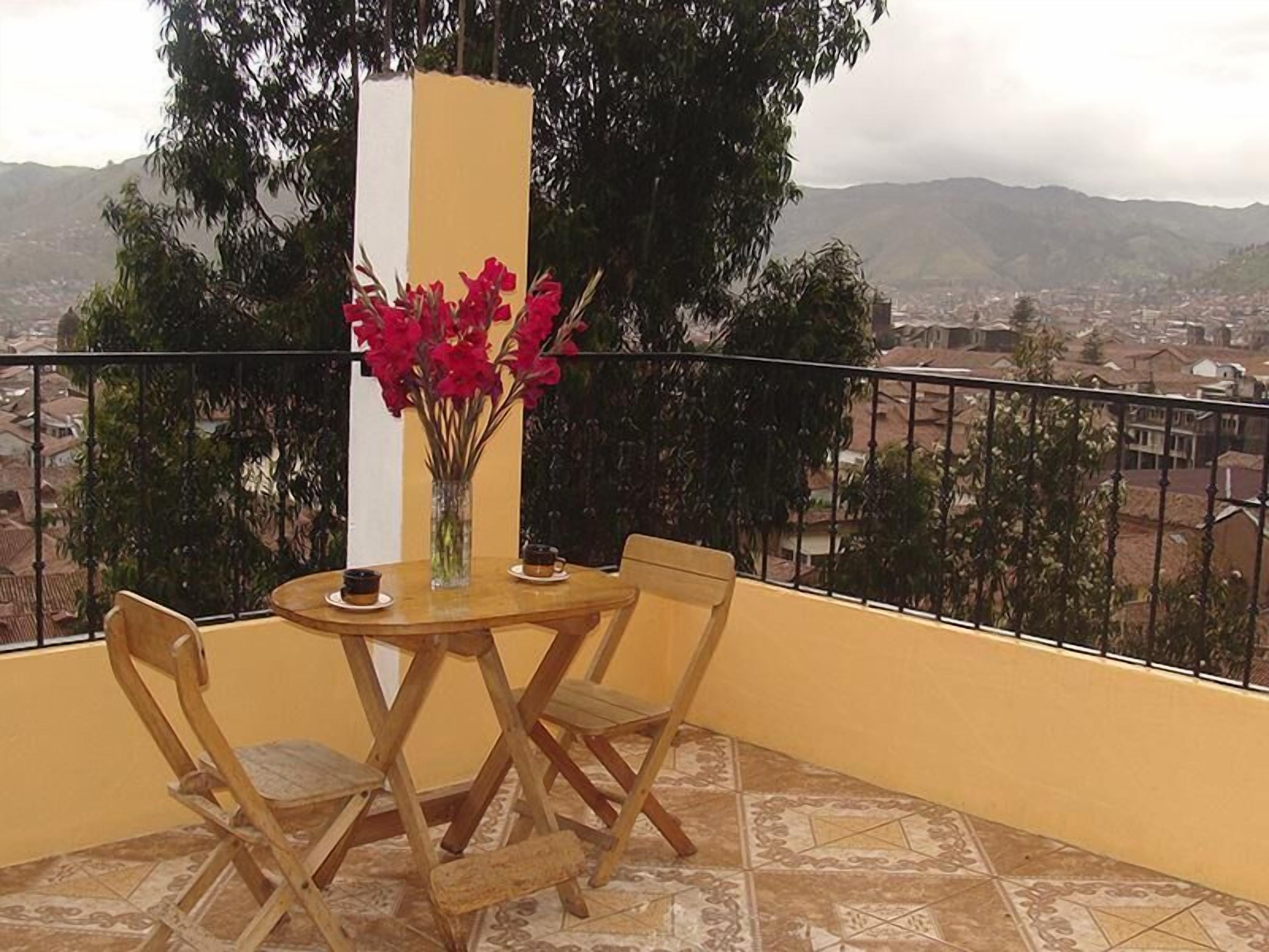 A balcony with a round wooden table, two chairs, a vase with red flowers, and a view of a cityscape with mountains in the background.