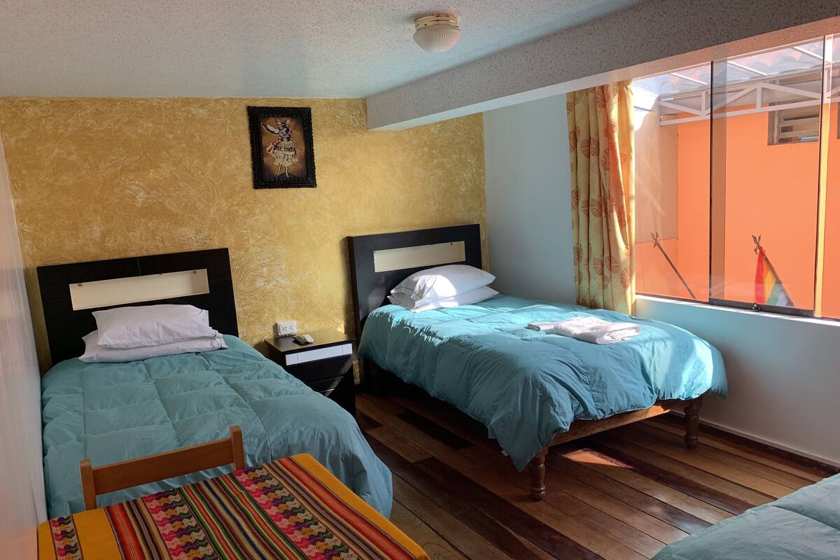 A room with two beds, a wooden floor, a small table with a colorful tablecloth, and a window with curtains.